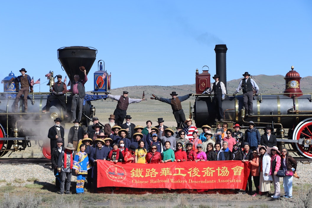 A group of people recreate a historic photograph, standing in front of two steam train engines