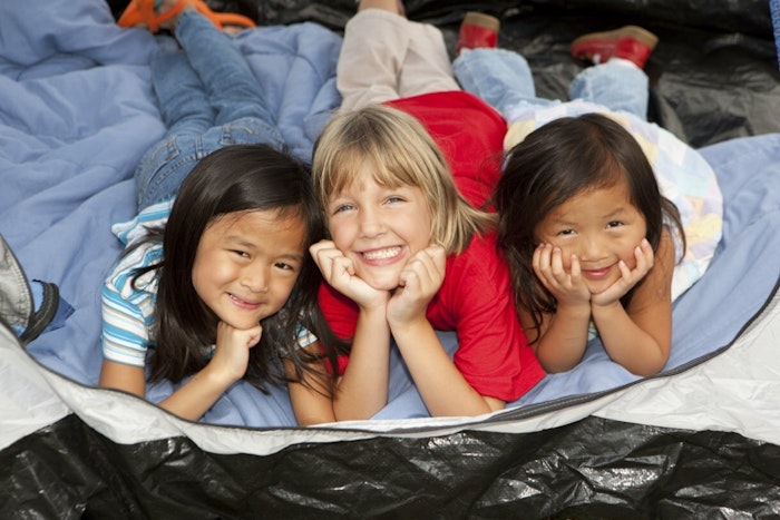 Three kids lay on their stomachs in a tent and propped up on their elbows smile at the camera