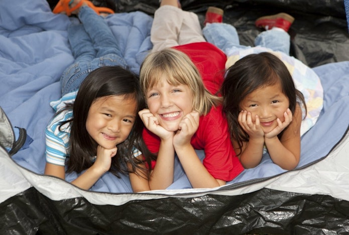 Three kids lay on their stomachs in a tent and propped up on their elbows smile at the camera