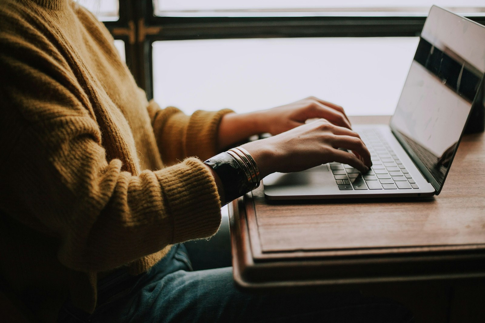 Close up of a person wearing a sweater and bracelets typing on a laptop
