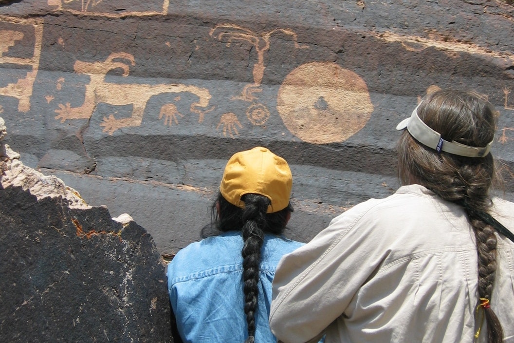 Two people, both with long braided hair, look at petroglyphs on stone