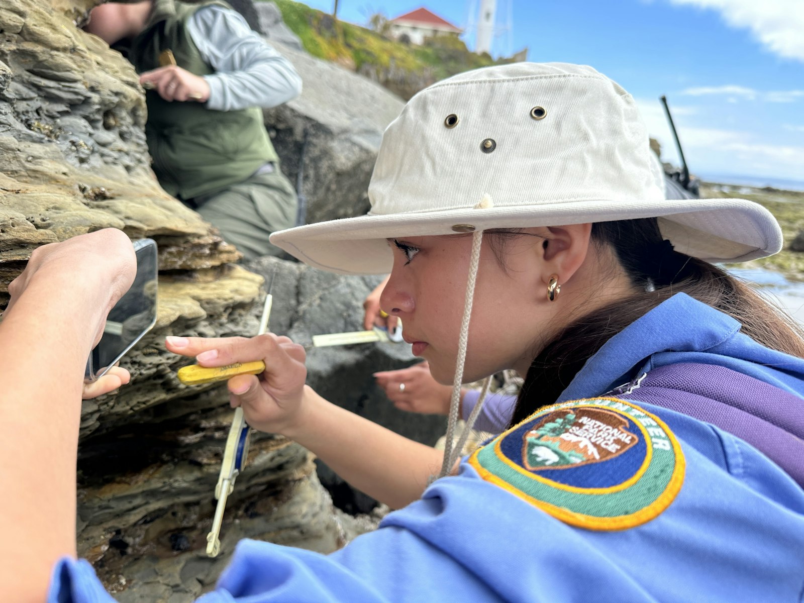 A girl wearing a fishing hat and a National Park Service Volunteer patch is inspecting rocks along the shore with a phone.