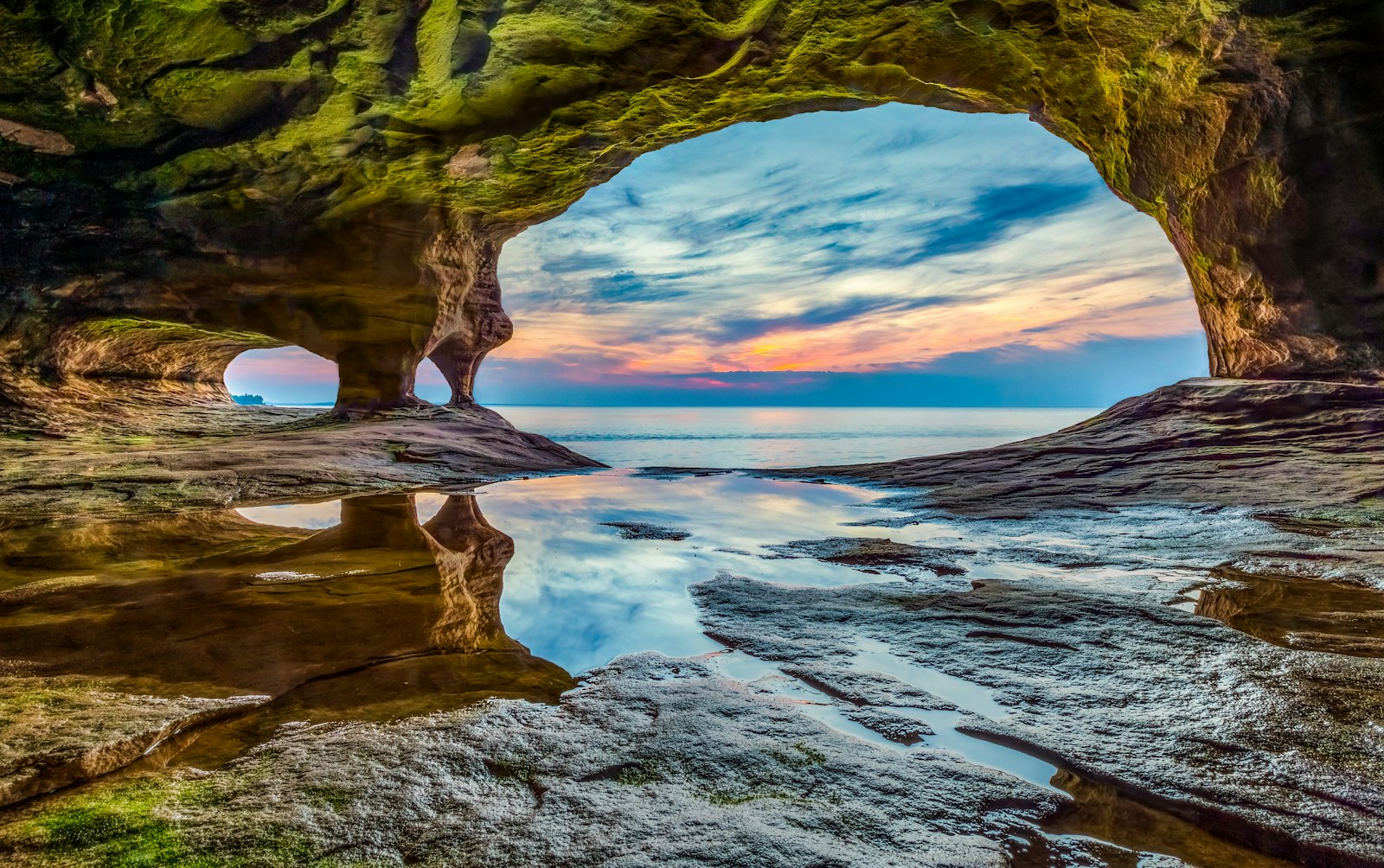 A rainbow sky with clouds is seen through the opening of a natural rock archway. In the foreground, a still lake pools into a rocky shore
