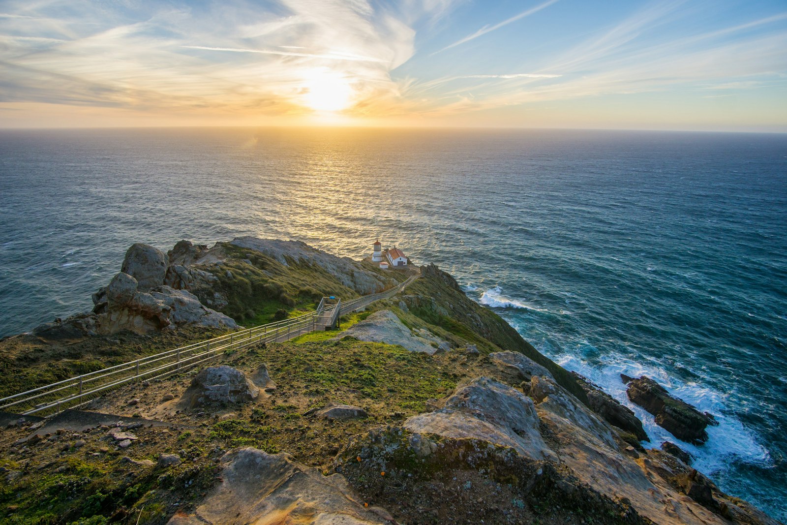 Four white-sided, red-roofed structures sit on a rocky headland above the Pacific Ocean at the base of a long stairway. Above and to the left of the lighthouse, the sun filters through wispy clouds as it descends toward the horizon.