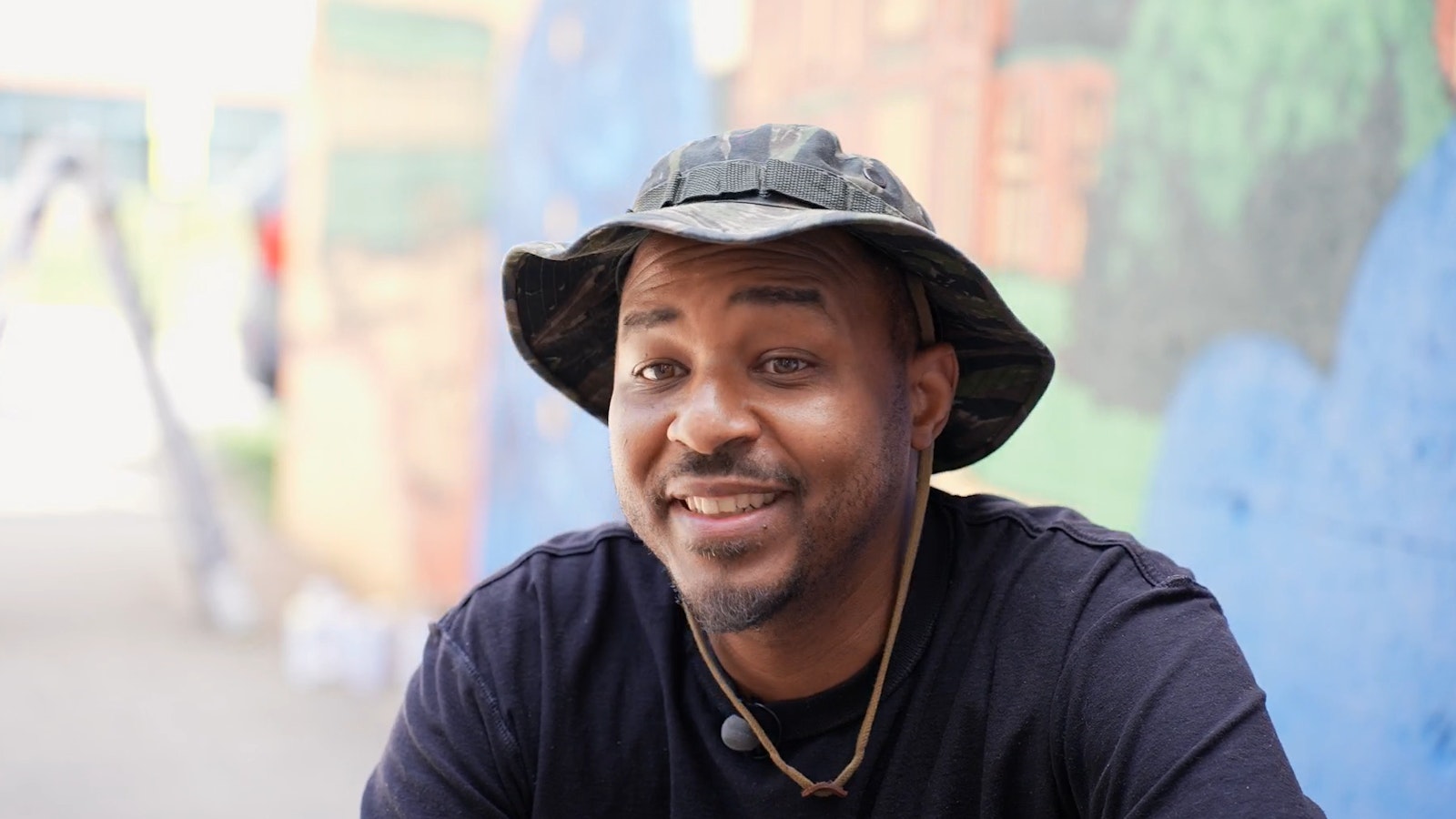 Artist Joe Nelson wears a black t-shirt and bucket hat, sits in front of a mural