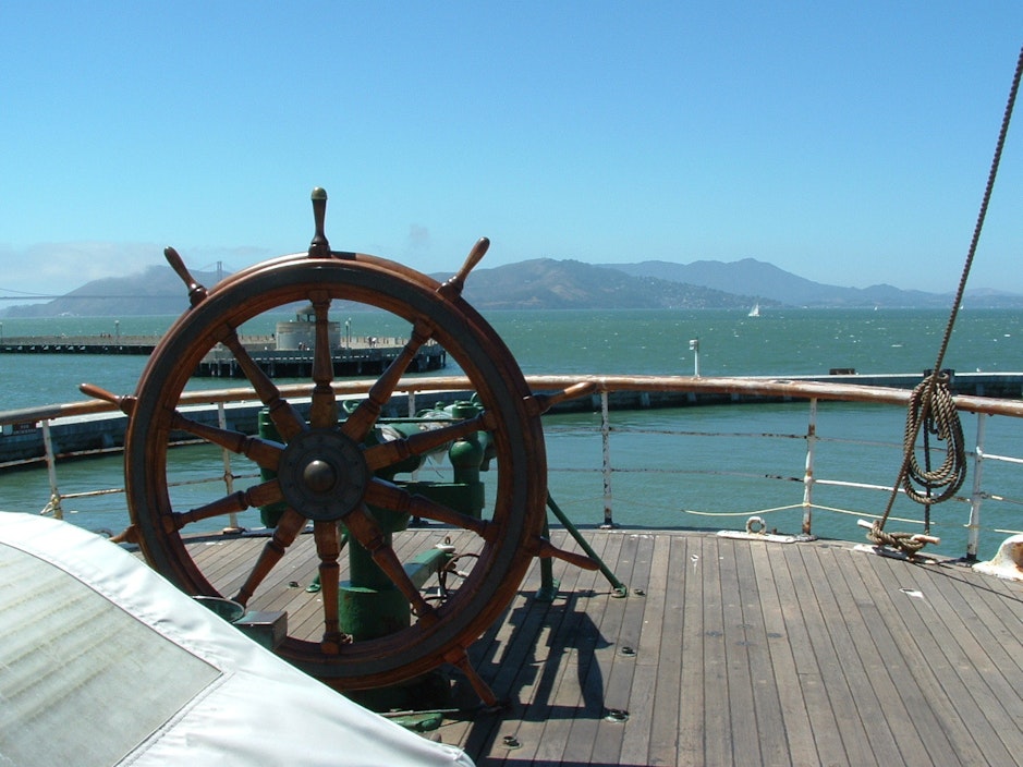 View from the deck of a boat, including a large wooden steering wheel and a deck guard rail