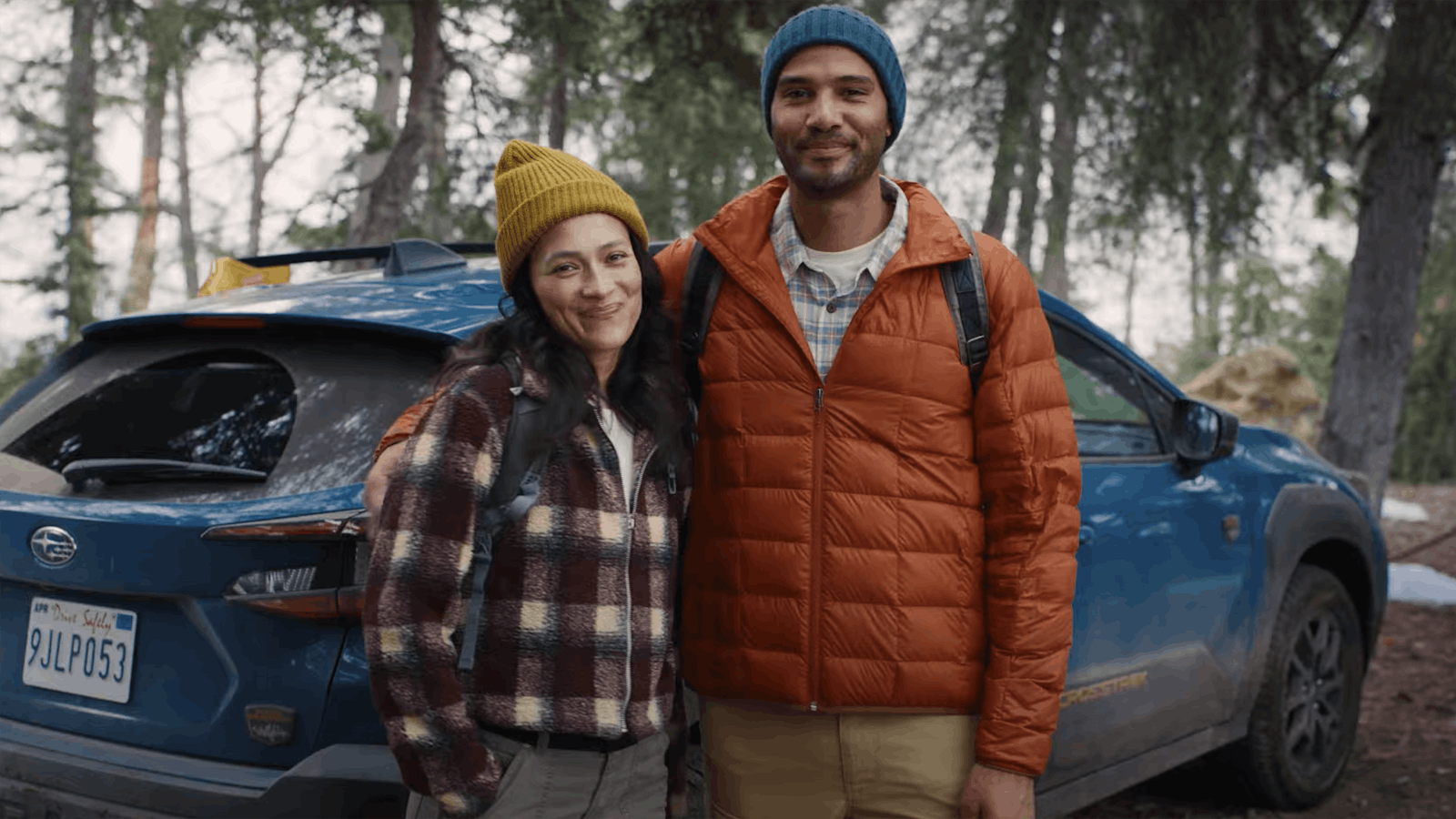 Two people stand in front of a car in a forest and smile for a camera
