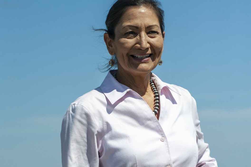 Woman wearing pale pink blouse smiles wide with blue sky and vast ocean behind her.