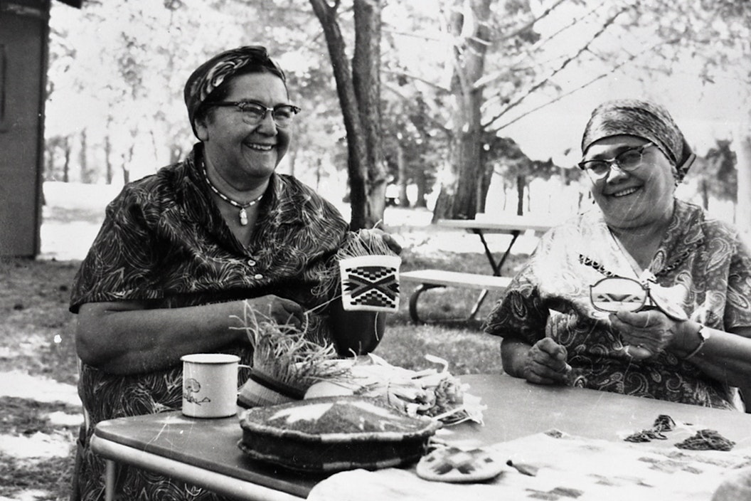 Vintage photograph of two women smiling as they demonstrate beading and cornhusk weaving