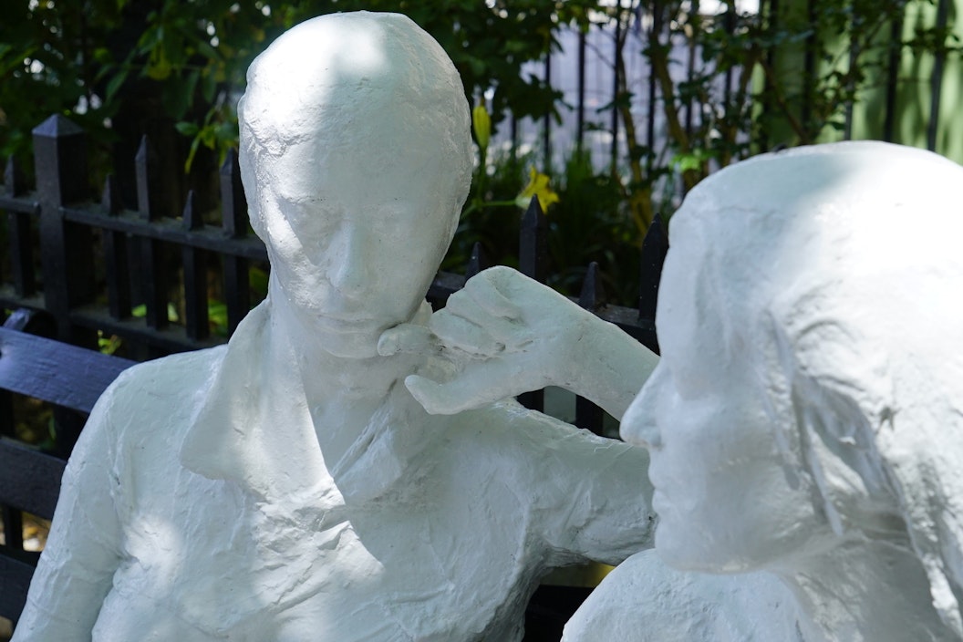 Two white plaster and stone sculptures depict two women sitting on a bench having a conversation