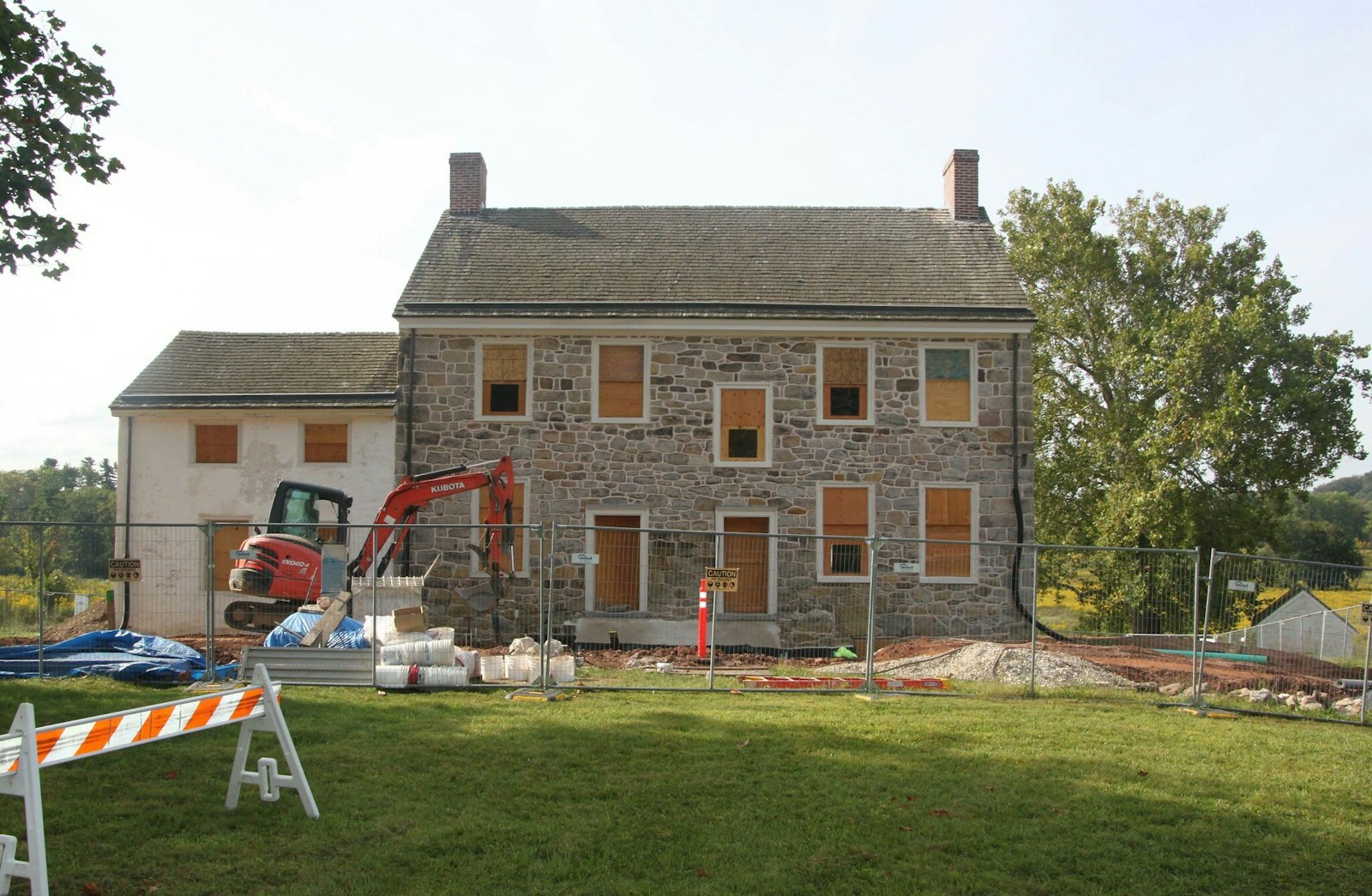 Construction equipment outside a two-story stone home with windows, surrounded by trees