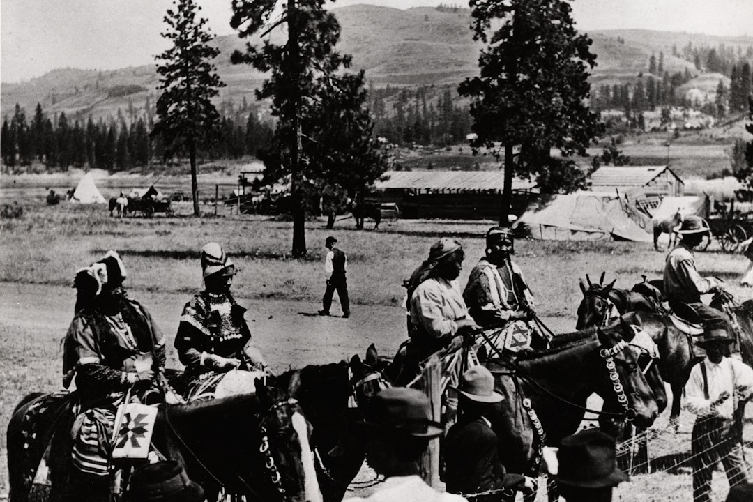 Black and white photograph of four women in traditional dress on horseback. In the background are rolling hills, pine trees, and several buildings and teepees.