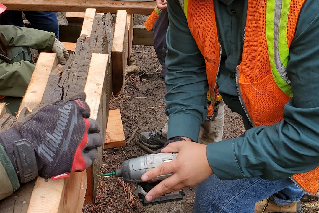 A person wearing a high-visibility vest and hard hat uses an electric drill to drill into the side of planks of wood.