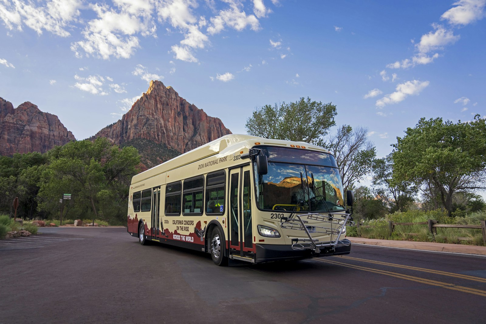 A beige shuttle bus on a dark asphalt road in front of a large, triangular sandstone mountain.