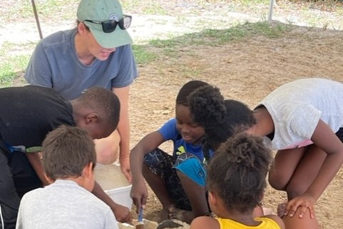 A group of children gather around the a dig site, accompanied by an adult wearing a baseball hat. They point at things and dig into the ground.