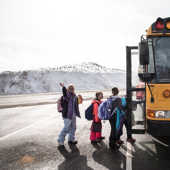 A group of children climb onto a school bus. One waves at the camera. In the distance, a snow-covered mountain range