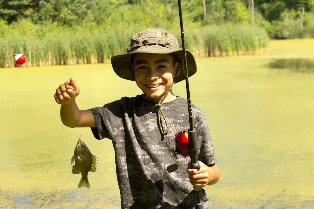 A child, wearing a bucket hat and holding a fishing rod in one hand, holds up a fish and smiles