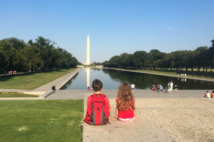 Two children sit and look away, across a long reflecting pool towards the Washington Monument