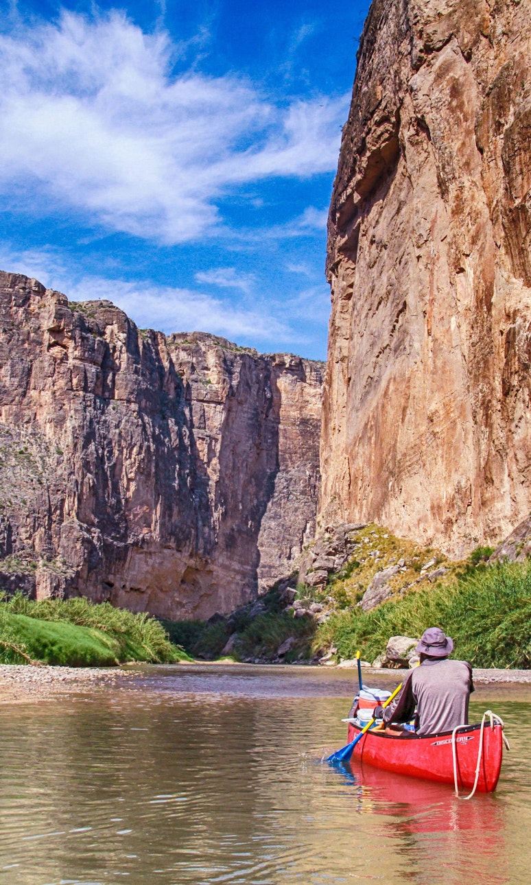 Two people kayak along a river at the base of a large canyon
