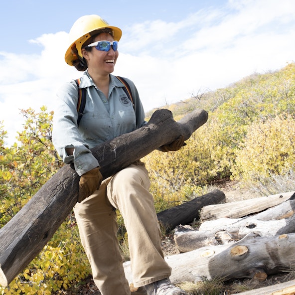 A service corps crew member, wearing a hard hat and sunglasses, carries a large log
