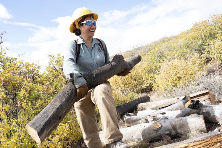 A service corps crew member, wearing a hard hat and sunglasses, carries a large log