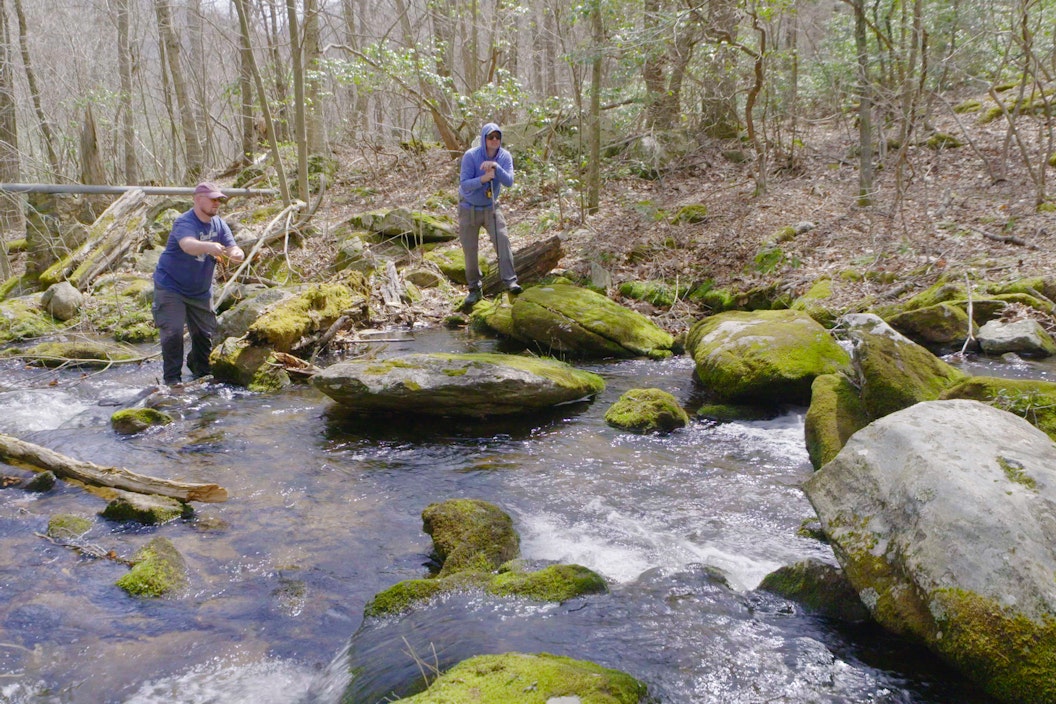 Two people fly fish along a stony river