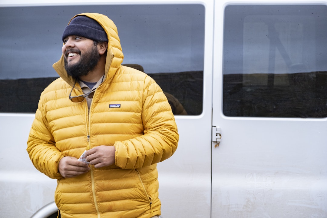 A person stands against a white van, wearing a puffy yellow jacket, and smiles