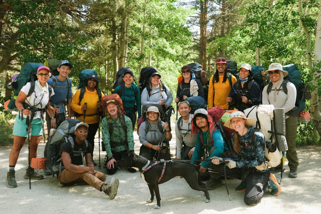 A group of a dozen people pose for a photo, all wearing hiking gear. In the foreground is a dog