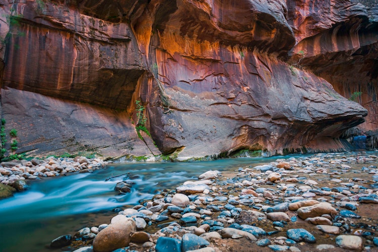 A stream runs over smooth, multi-colored stones at the side of a red canyon wall
