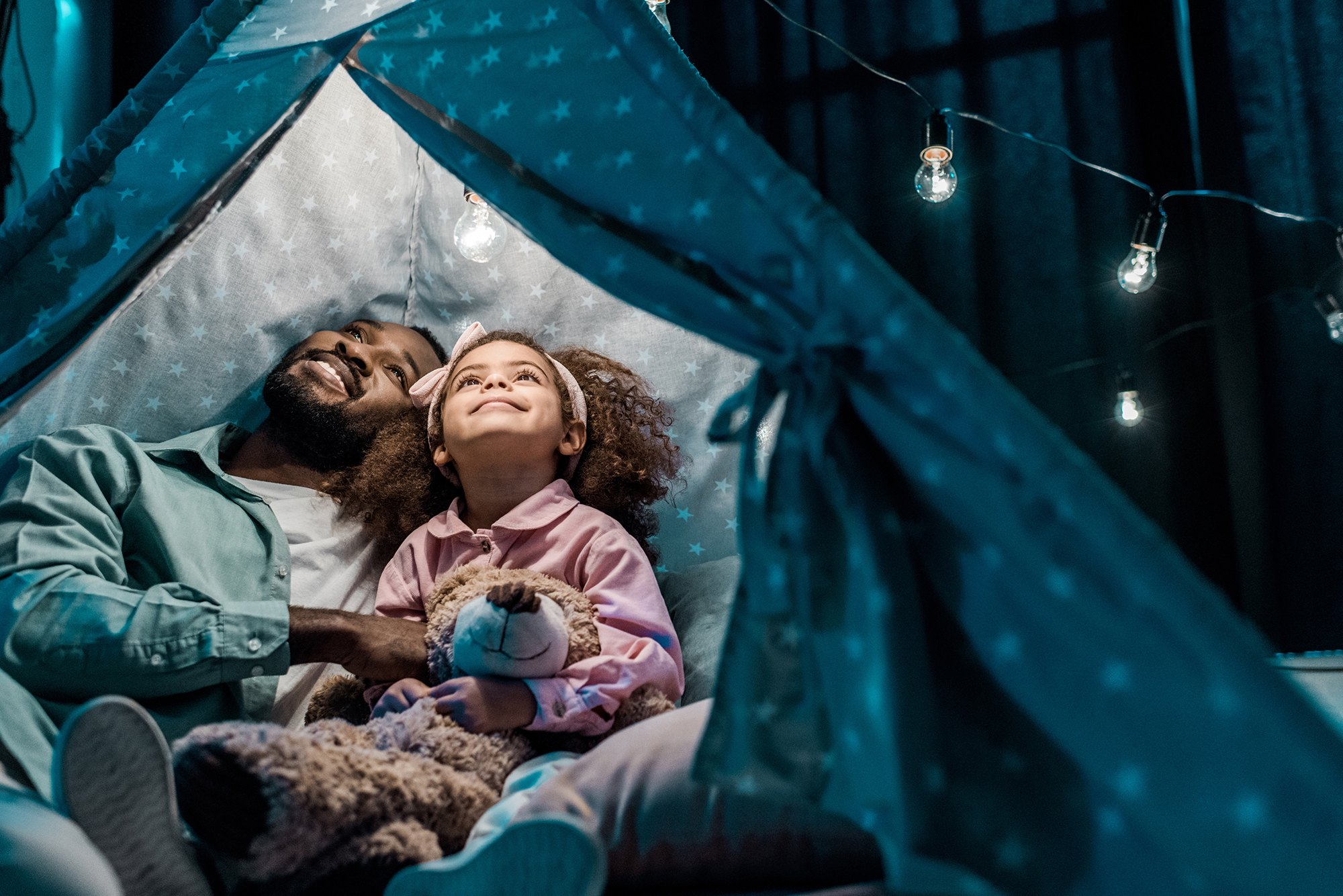 Family inside a tent pitched indoors, looking up towards strings of light