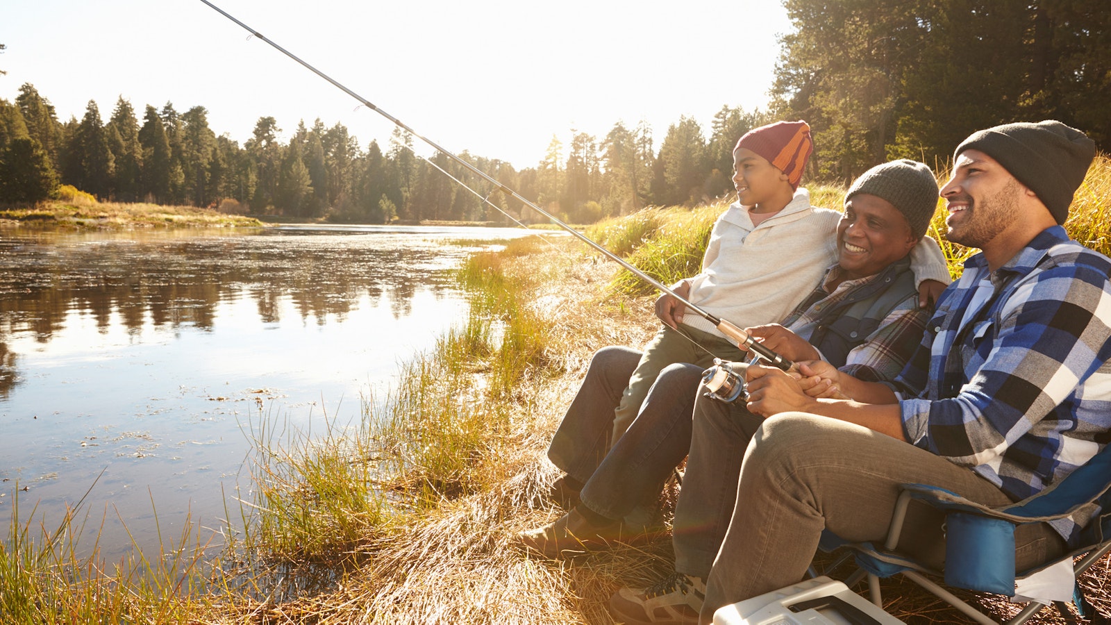 Three people sit on a riverbank and fish