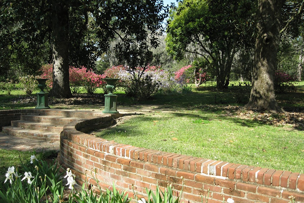 Winding paved path, lined with brick retaining walls, leads through a blooming garden capped off with shady trees